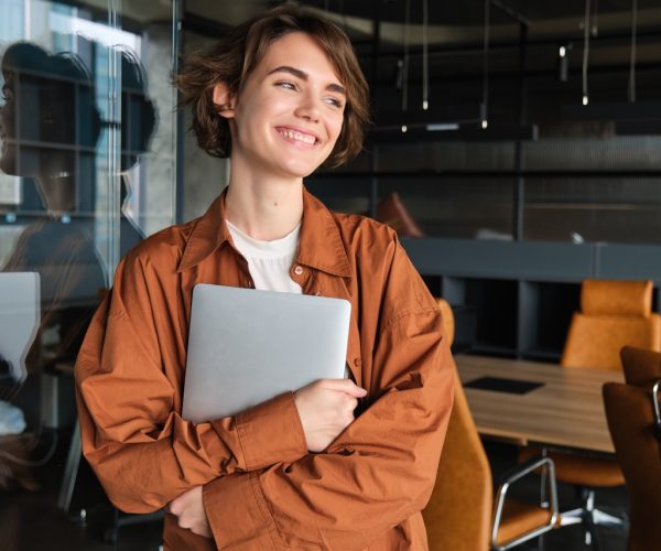Portrait of young female manager, office worker with laptop, digital nomad programmer working at her workplace, standing with computer and smiling.