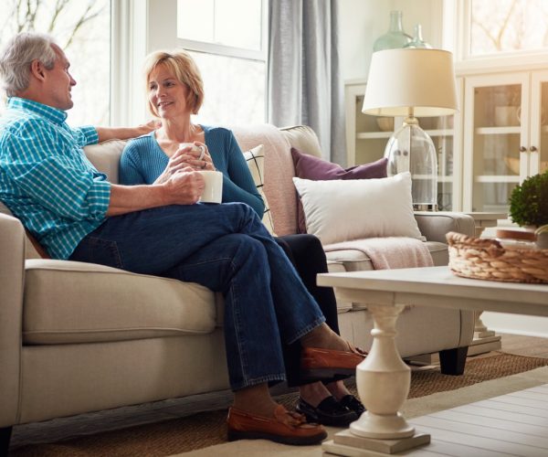 Senior couple, coffee and smile on sofa in conversation, relax or happy memory in home living room. Old man, elderly woman and chat for bonding with tea, love or story in retirement on couch in house.